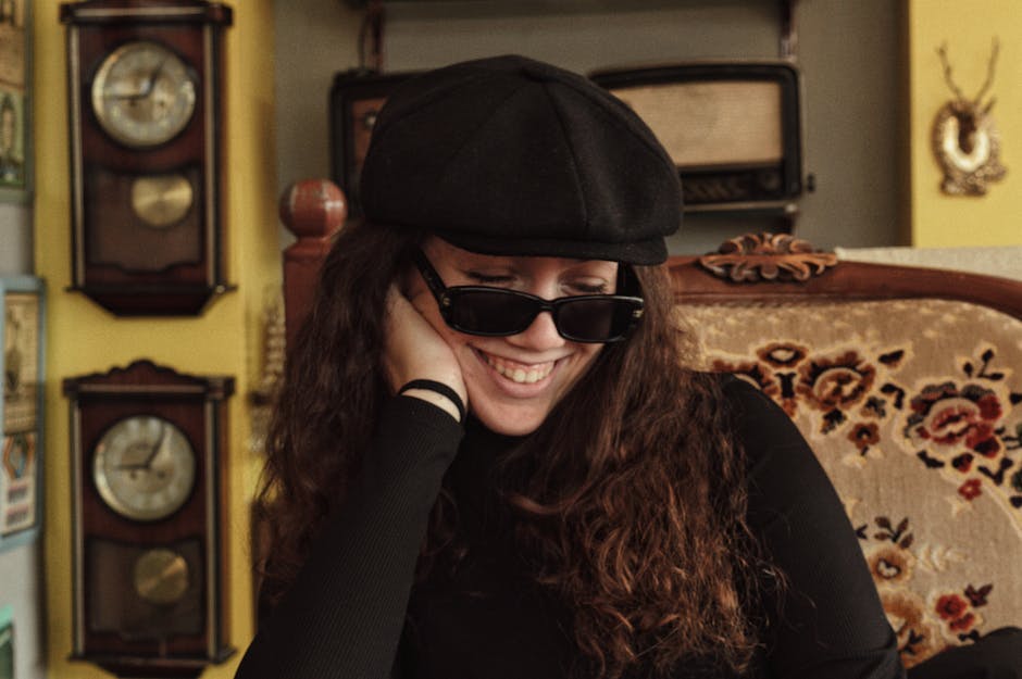 A woman wearing sunglasses and a cap smiles joyfully in a vintage-styled room.