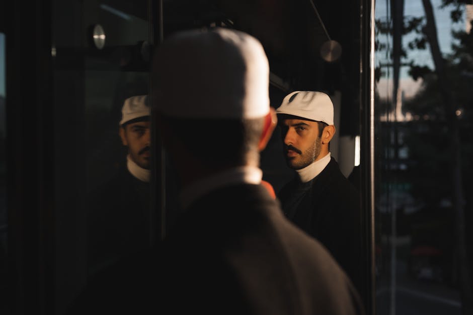 A man in a white cap stares at his reflection on a city street at dusk.