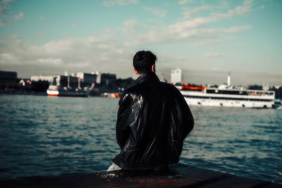 A man in a leather jacket sits by the harbor, gazing at the calm waters under a clear sky.