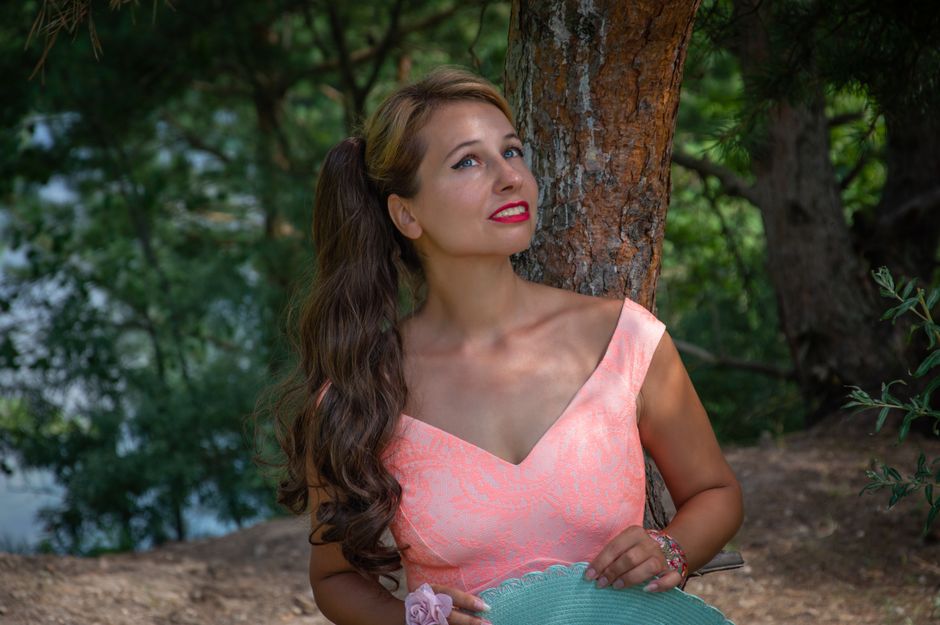 A woman with long hair and pink dress enjoying the outdoors next to a tree.