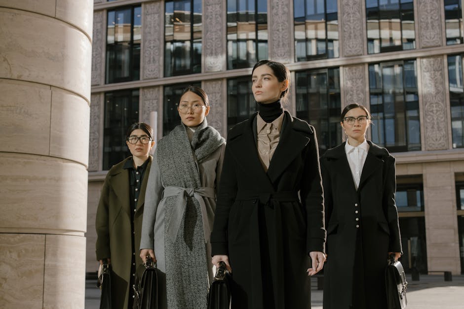 Four confident women in coats with briefcases outside an urban office building.